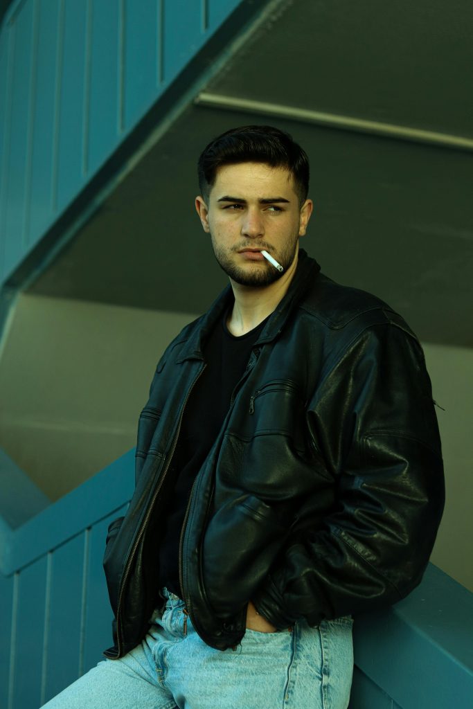 Portrait of a man in a black leather jacket smoking a cigarette against a blue background.