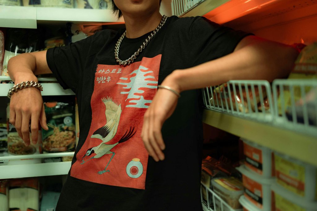 A stylish young man wearing a graphic T-shirt and jewelry poses casually in a grocery store aisle.