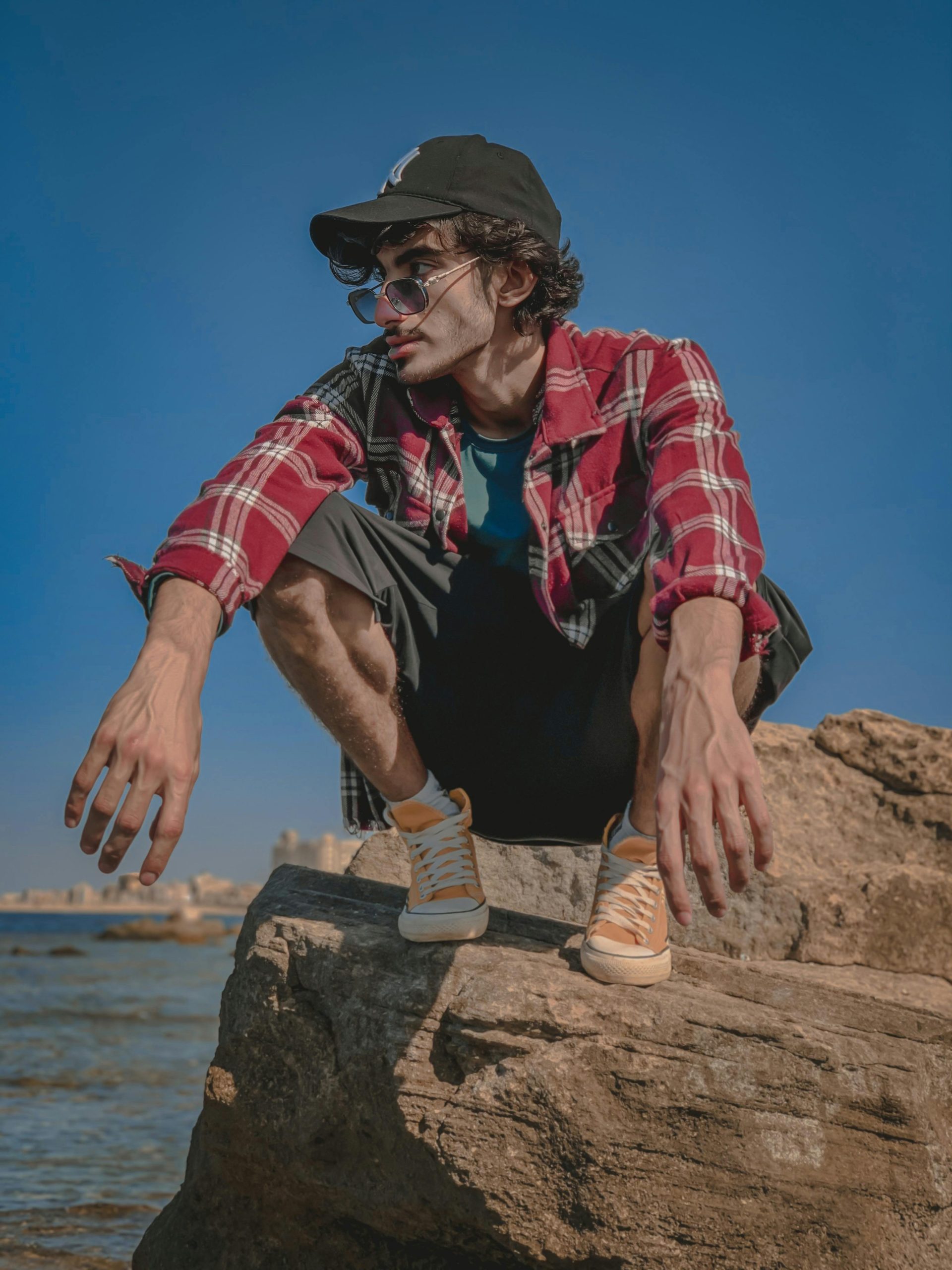 Young man in a red plaid shirt squatting on coastal rocks, showcasing a trendy casual fashion look.